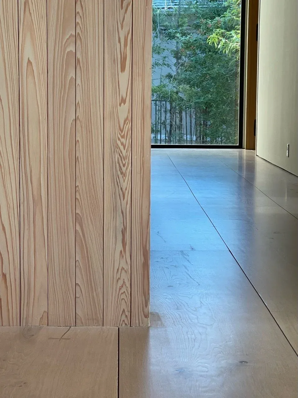 Interior detail showing wood panel wall and flooring with a view to exterior greenery through a large window.