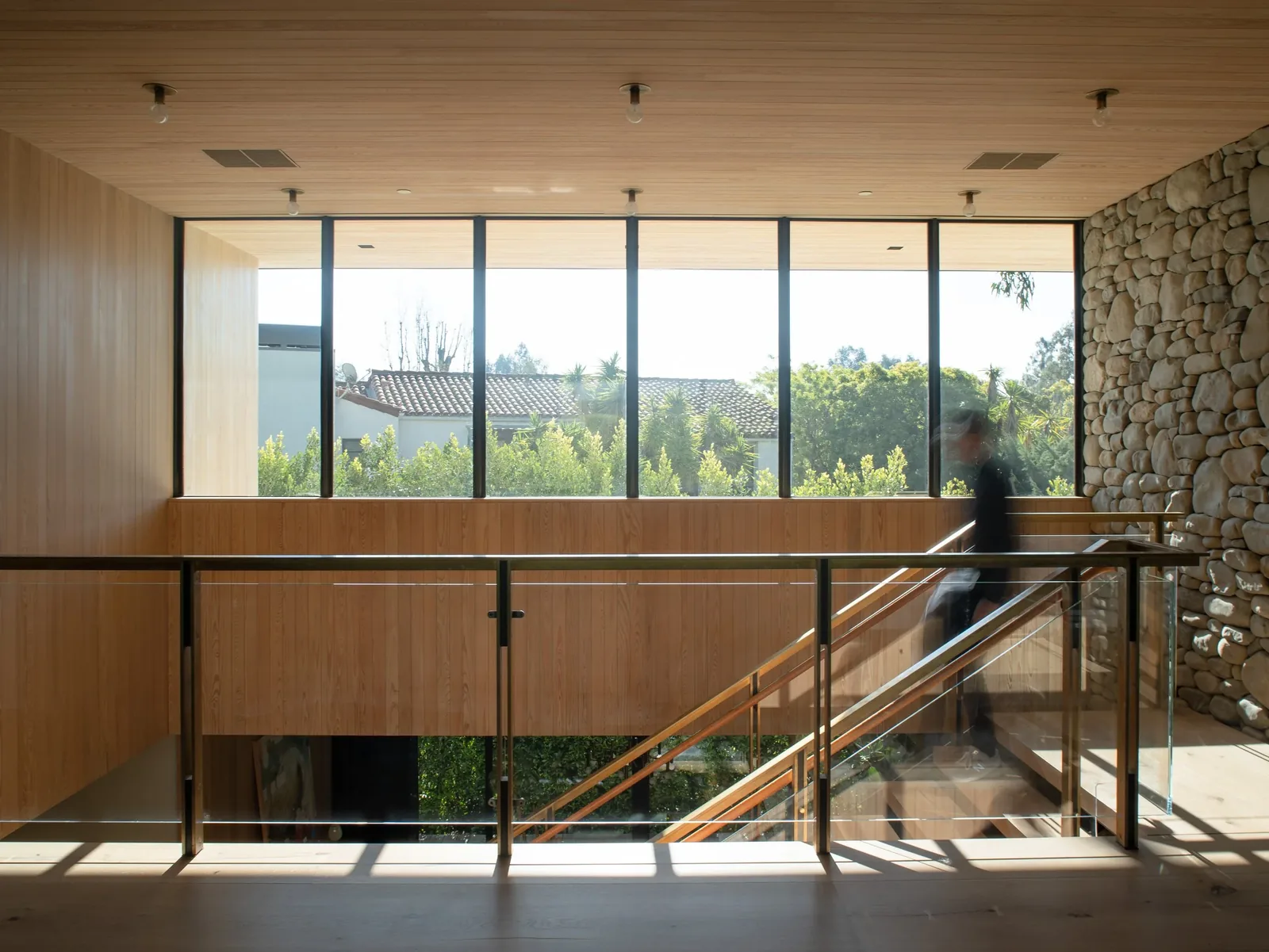 A wood-paneled stairwell with floor-to-ceiling windows, glass railing, and a stone accent wall.