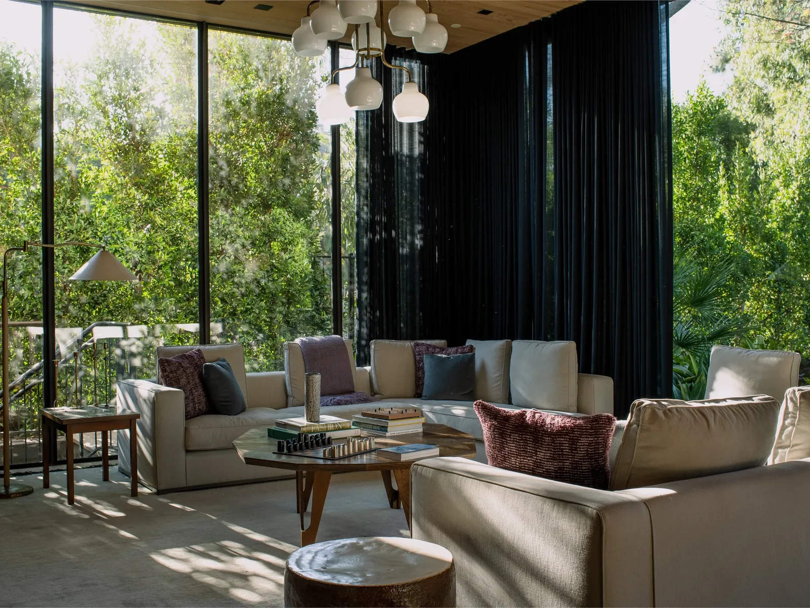 Living room with floor-to-ceiling glass walls overlooking trees and featuring white sofas and a central coffee table with books and decor.