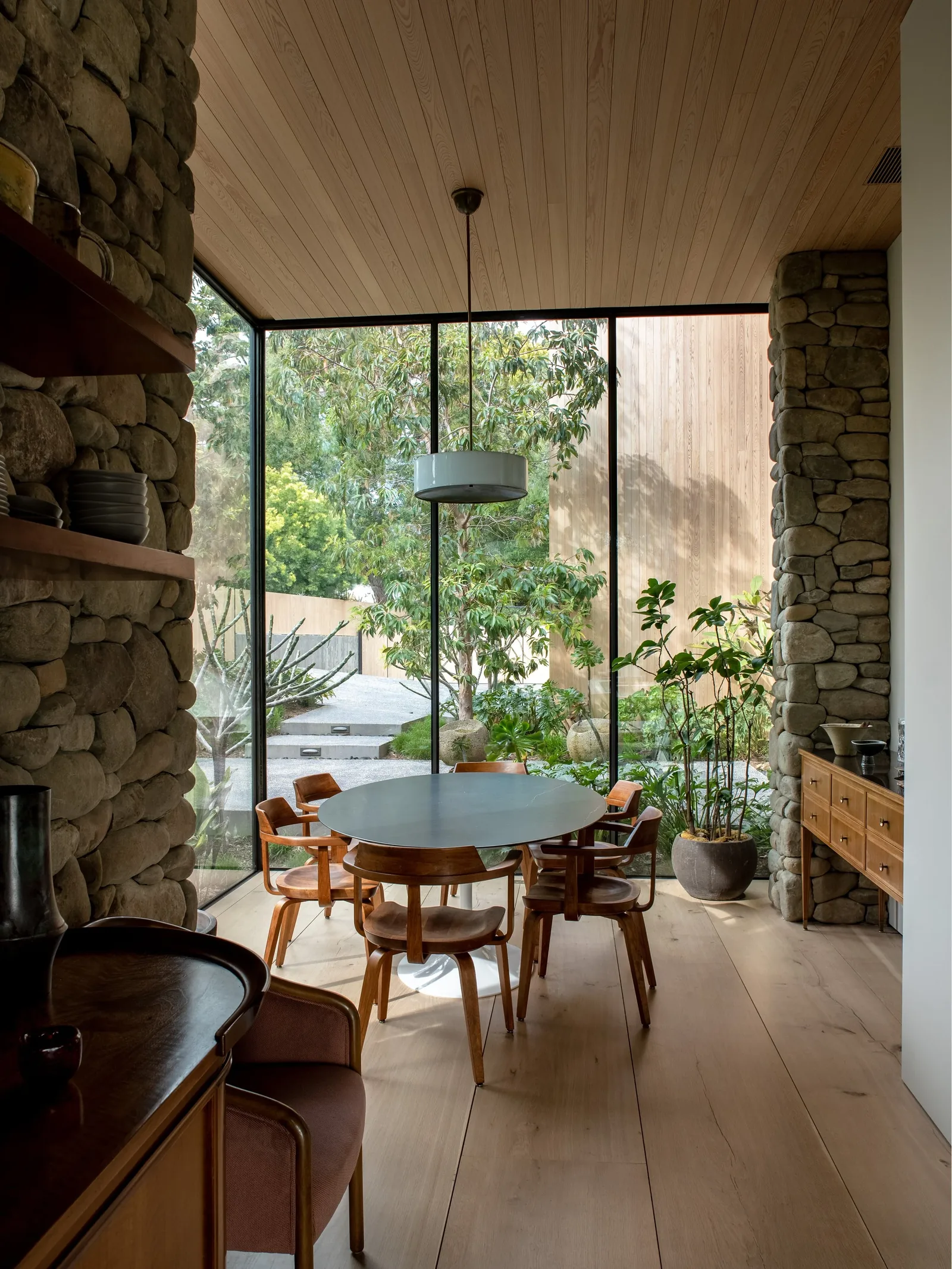 Dining area with wood-paneled ceiling and floor-to-ceiling glass windows next to stone walls.