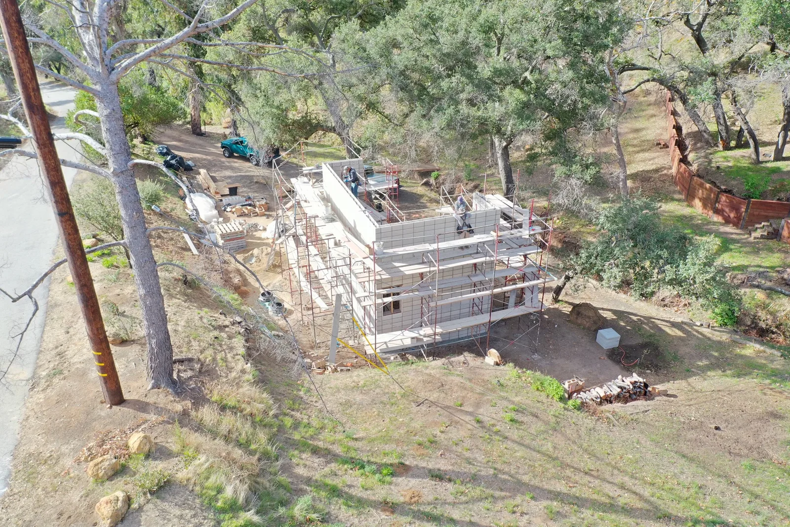 A residential building with block walls is under construction on a hillside surrounded by trees and construction materials.