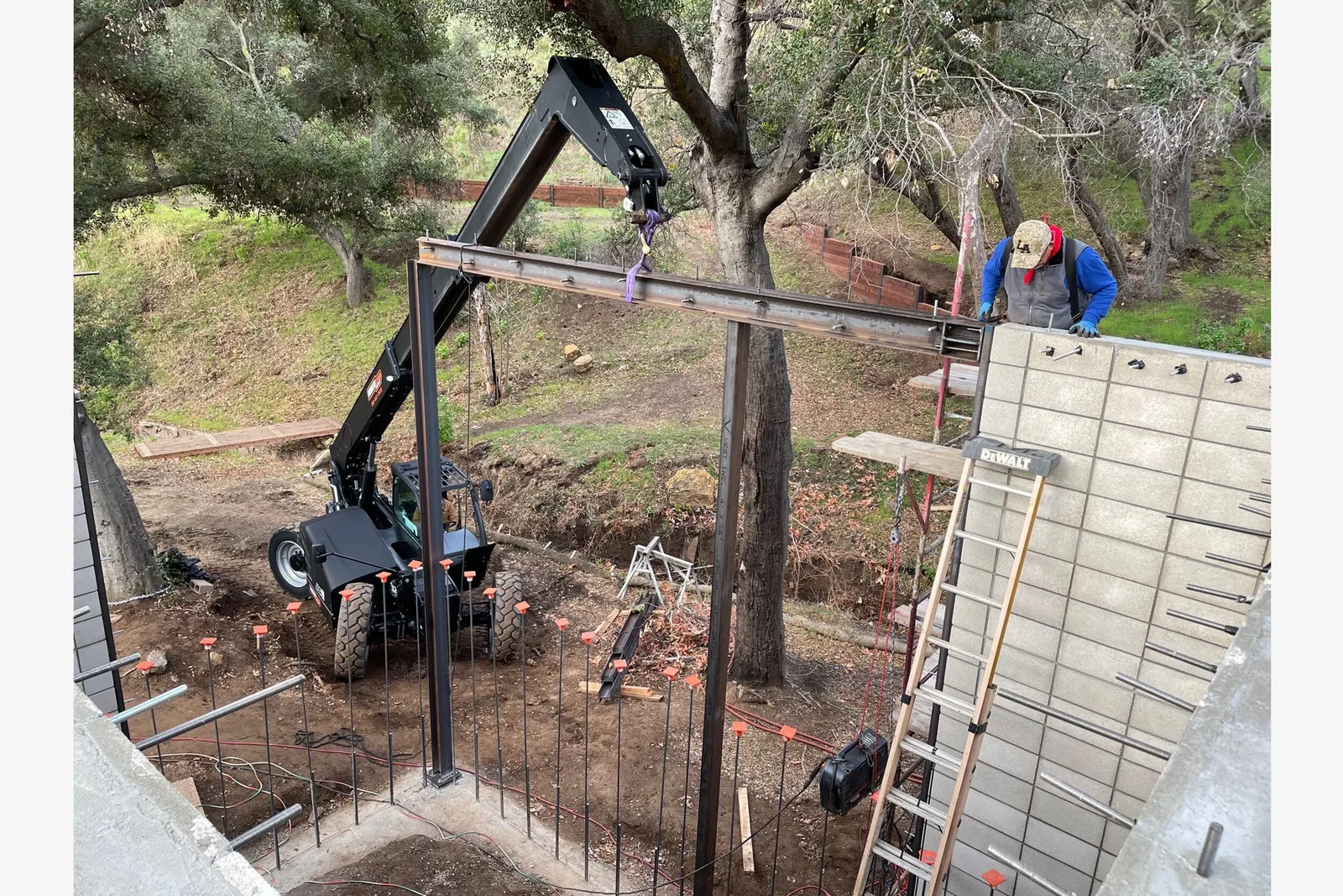 Steel framing is lifted at a construction site, with block wall foundation work visible beneath trees.