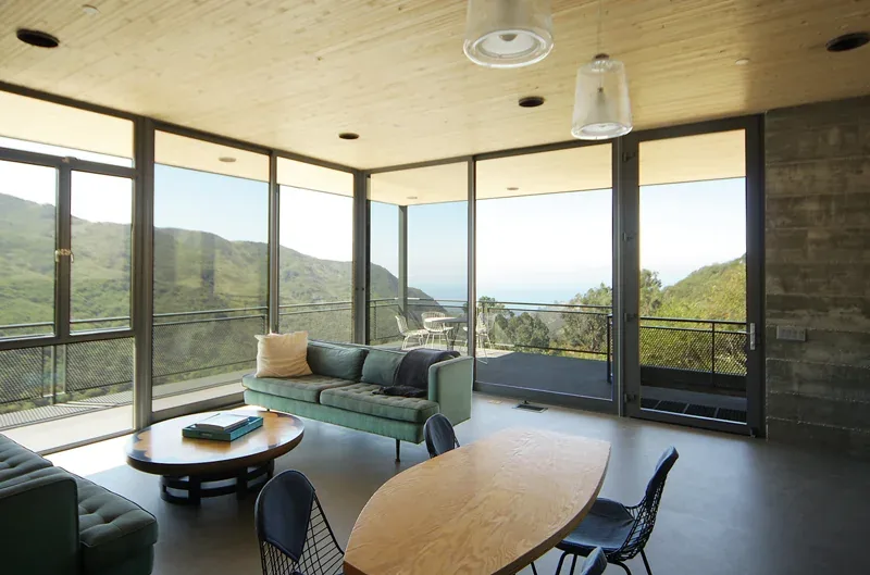 Living room with large glass walls and a balcony providing expansive views of hills and ocean.