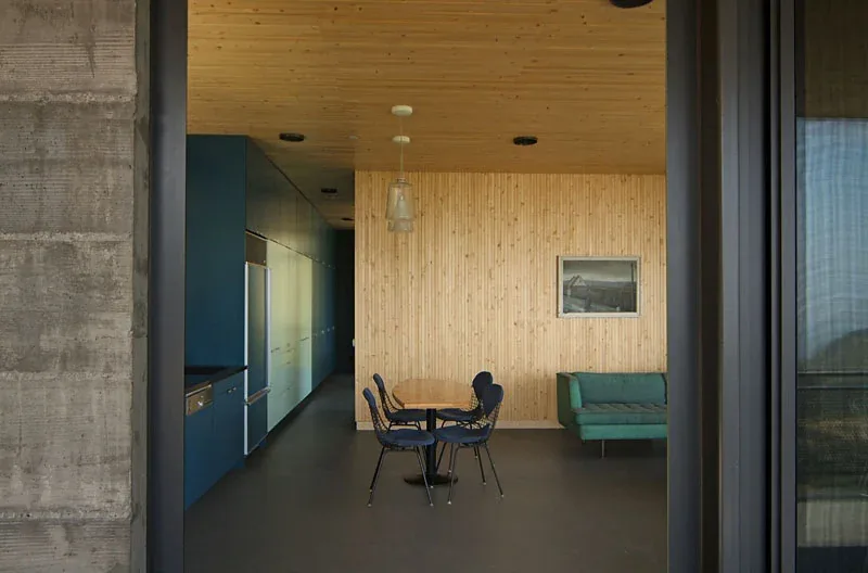 Modern kitchen and dining area feature wood-paneled ceiling and wall, blue cabinetry, and a concrete wall.