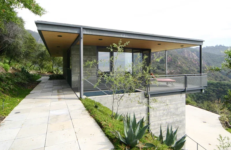 Exterior front view of a modern hillside house with board-formed concrete and extensive glazing under a flat roof with wood soffit.