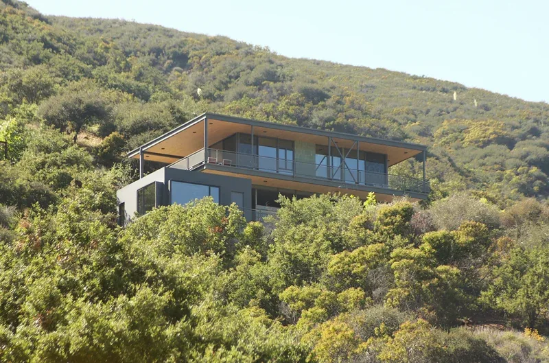 A modern hillside house with large windows and a covered balcony, nestled among dense greenery.