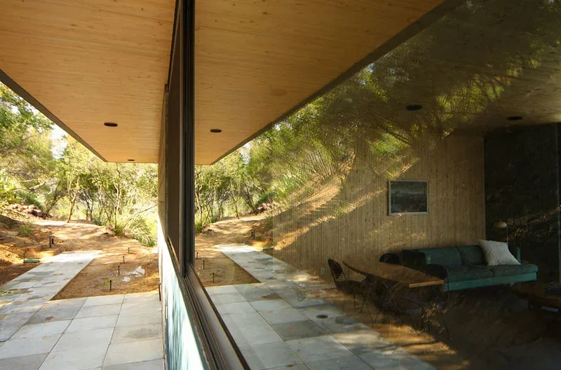 Large reflective glass window shows outdoor paved path and wood-textured exterior wall with seating area inside.