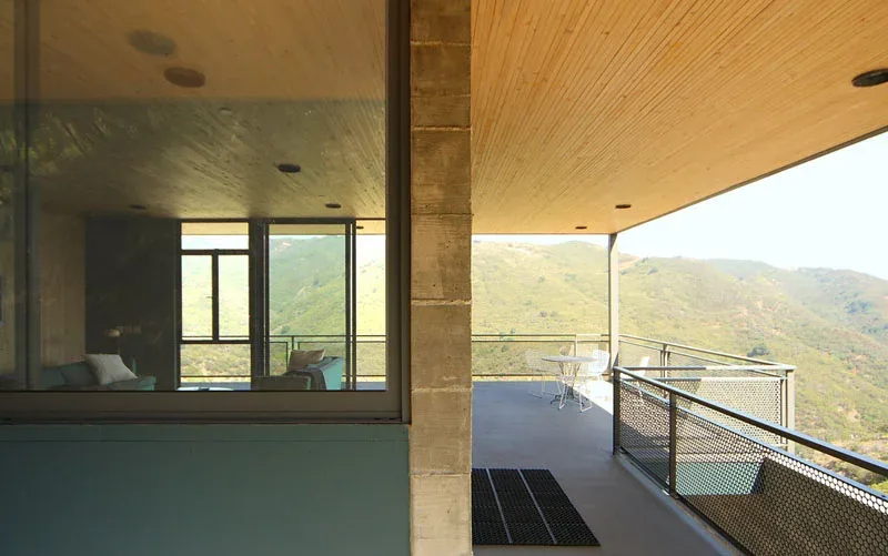Concrete balcony with metal mesh railing overlooking mountainous terrain, next to a floor-to-ceiling glazed room.