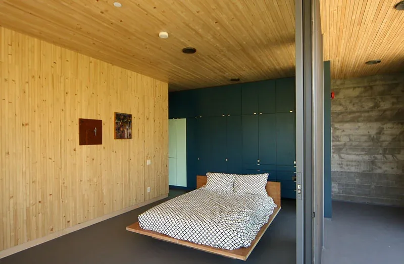 Interior bedroom with wood panel ceiling and wall, blue cabinetry, and concrete wall.