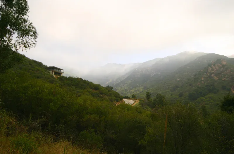 Exterior front view of a hillside house with natural mountainous surroundings under an overcast sky.