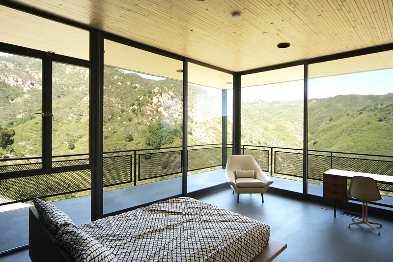 Bedroom with floor-to-ceiling glazing on two sides providing panoramic views of surrounding hills.