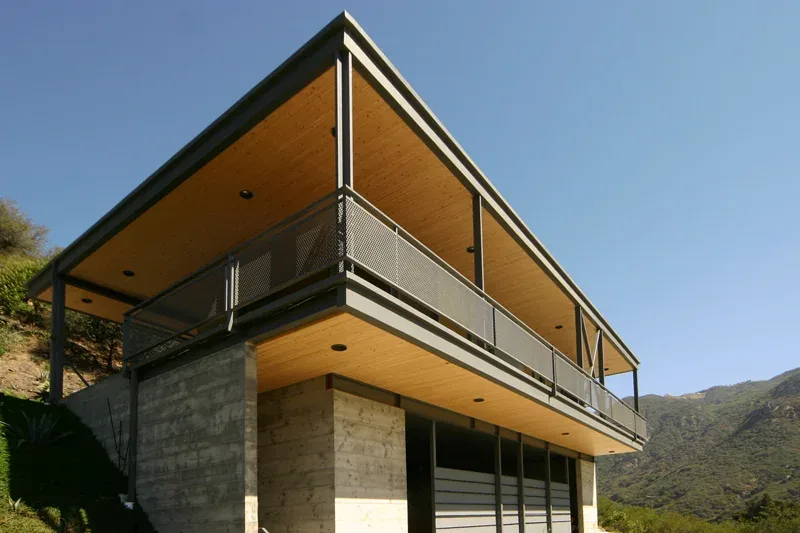 Modern two-story house with cantilevered upper floor, wood soffits, concrete base, and metal railings.