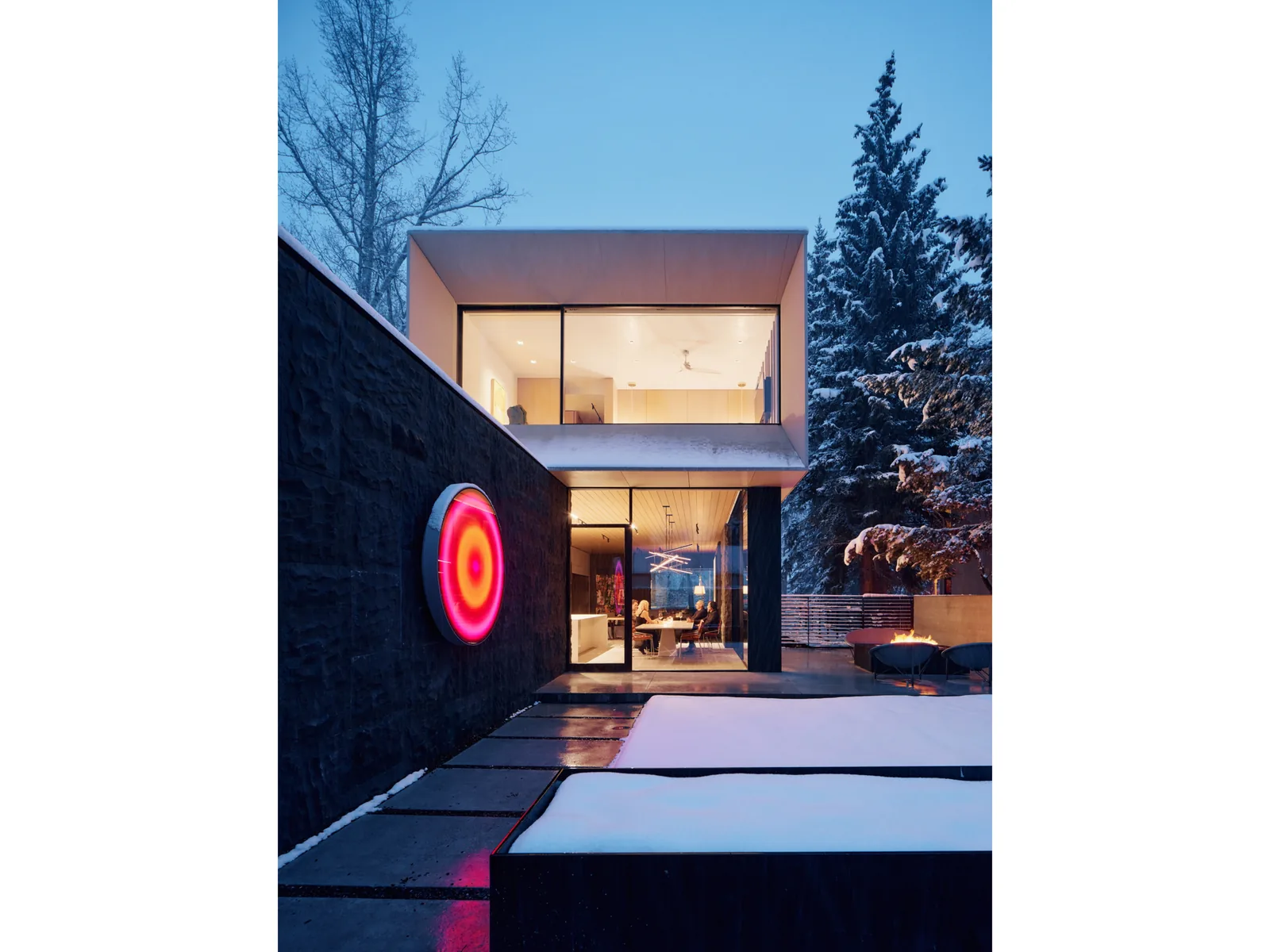 Two-story modern house with floor-to-ceiling glazing and dark stone exterior wall shown at dusk with snow-covered landscape.
