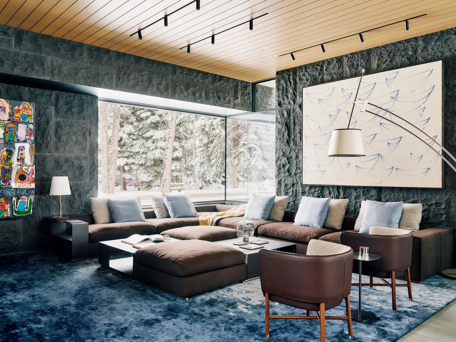 Living room featuring textured stone walls, corner floor-to-ceiling window, and a large sectional sofa around a central coffee table.