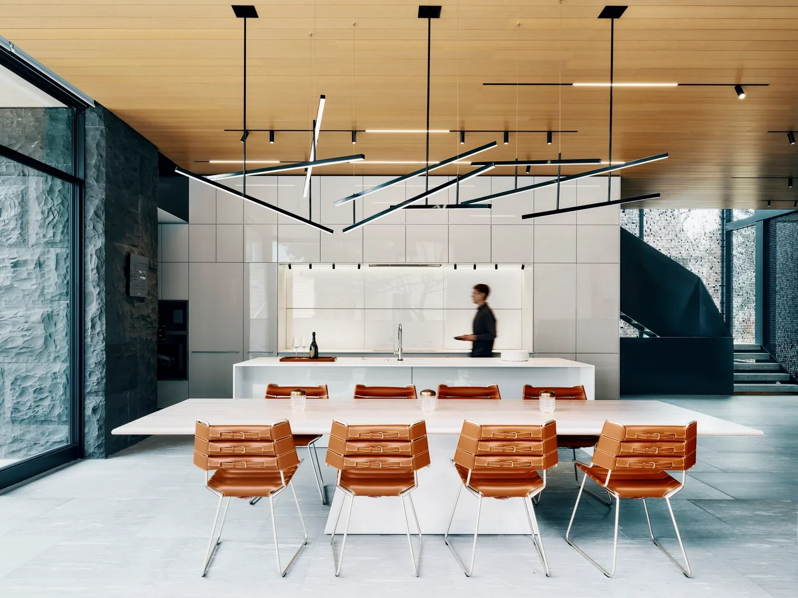 A view of the kitchen and dining area featuring a large white island, tan leather dining chairs, and linear pendant lights against a wood-paneled ceiling.