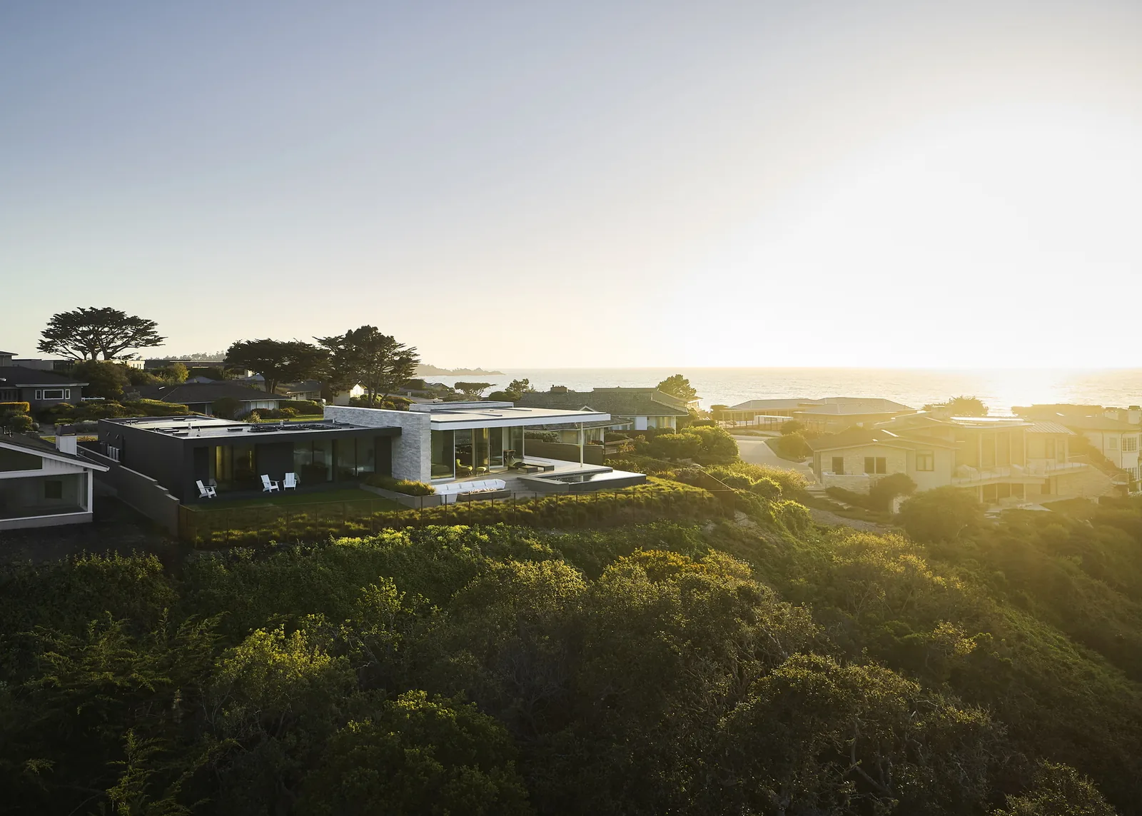 A modern hillside residence with flat roofs and large glass walls overlooking the ocean at sunset.