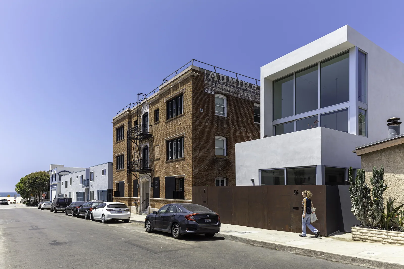 A modern cubic stucco building contrasts with a vintage brick apartment building on a coastal urban street.