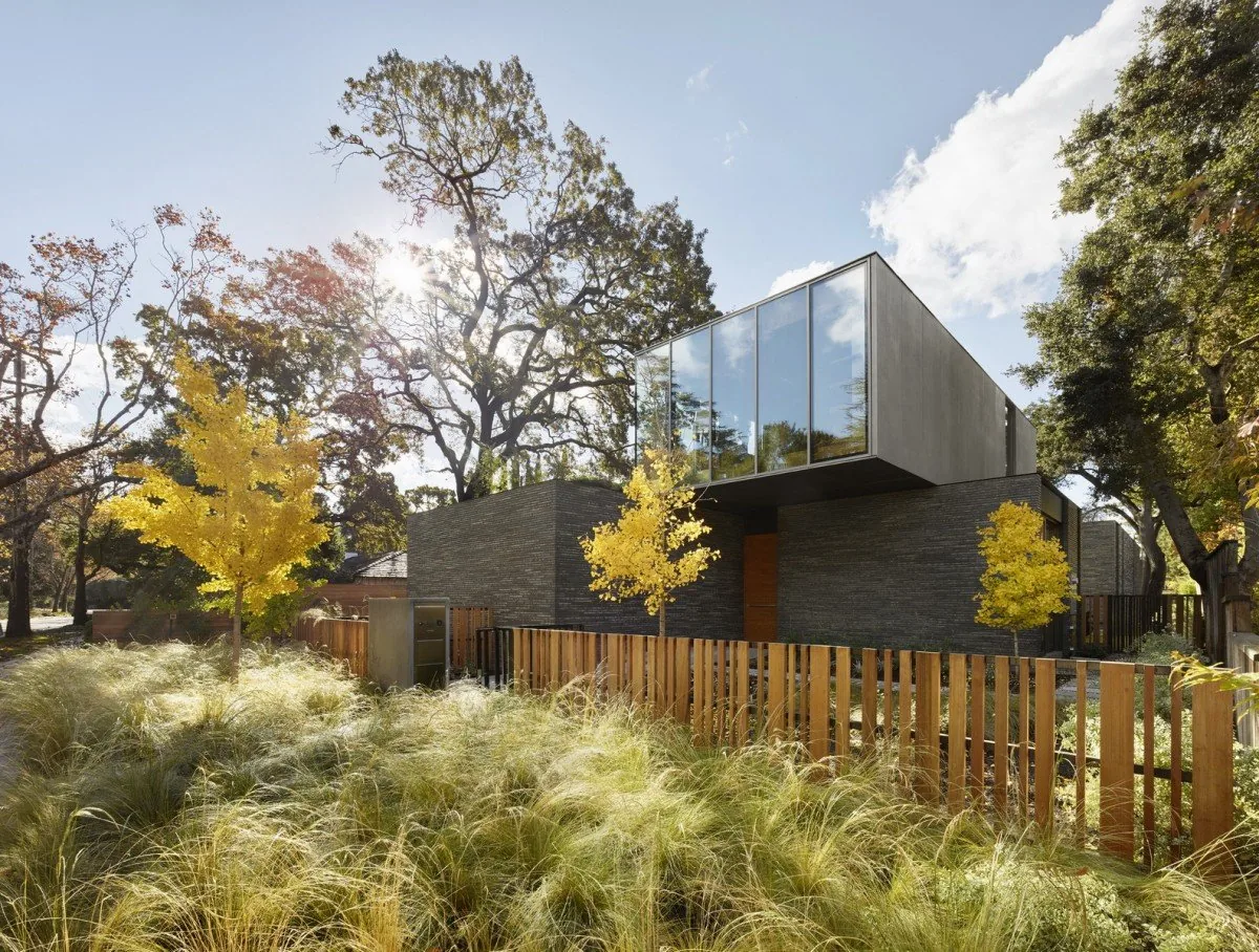 Exterior front view of a modern two-story house with floor-to-ceiling glazing, dark brick facade, and wooden fence.