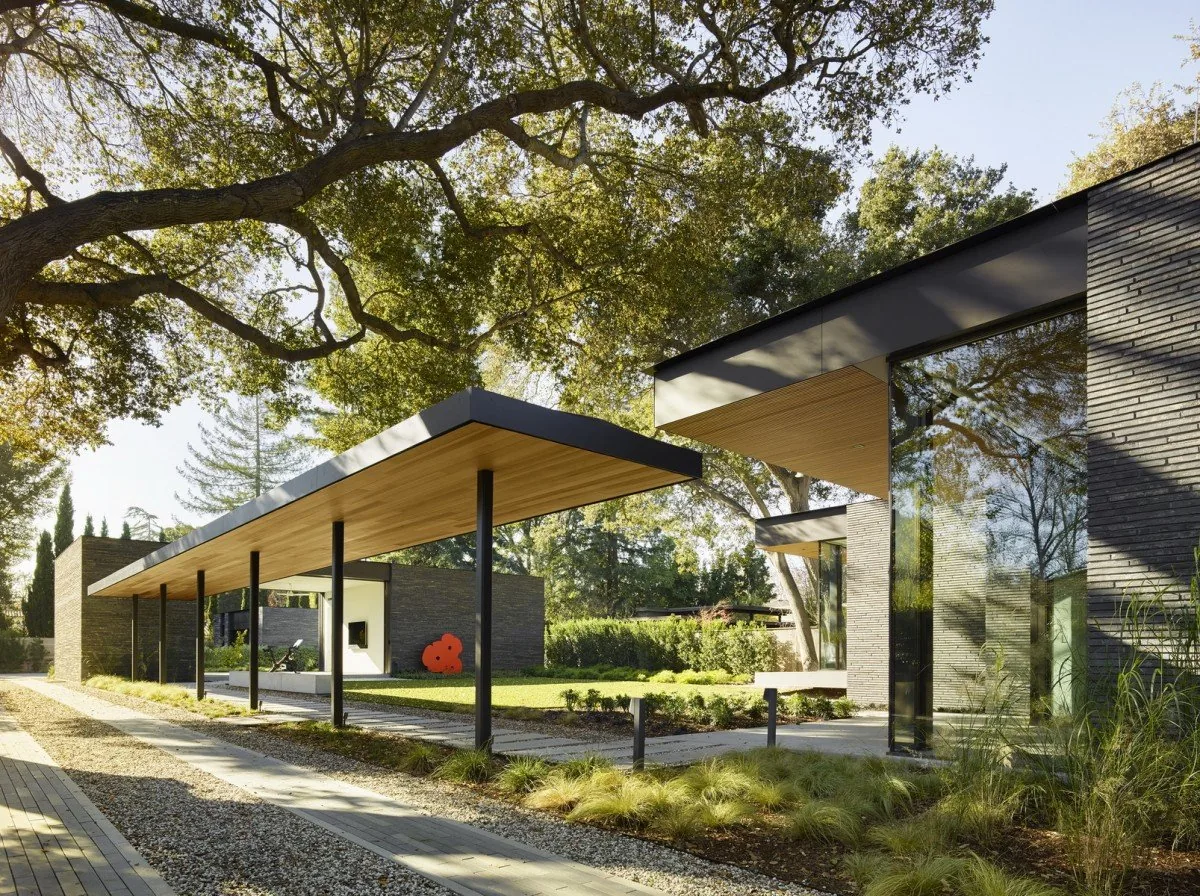 Exterior view of a modern house with flat roofs, wood soffits, large floor-to-ceiling glazing, and dark brick walls surrounded by landscaping.