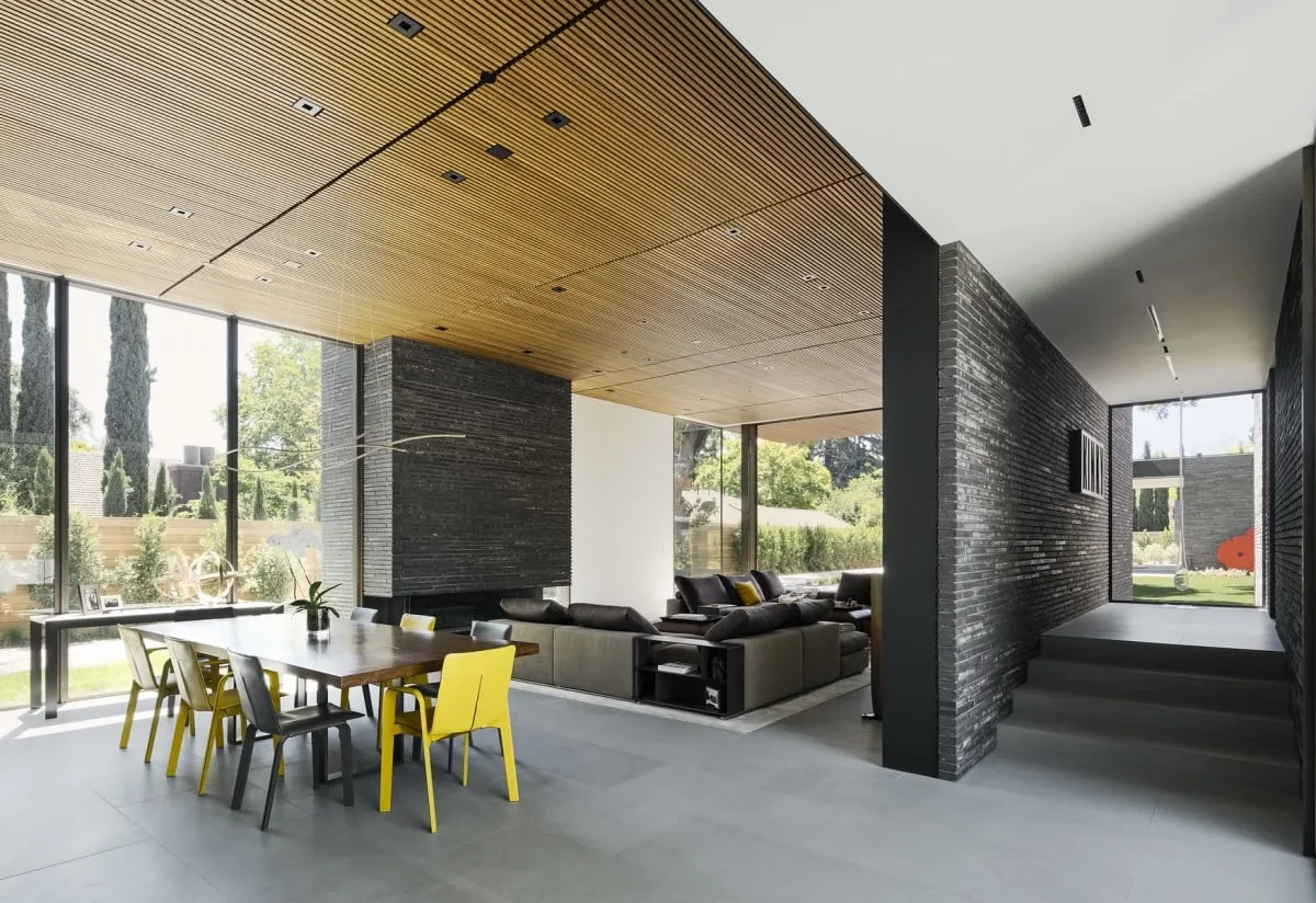 Modern living and dining space with wood slatted ceiling, gray brick fireplace, and large glass windows.