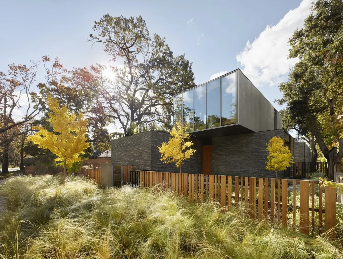 Exterior front view of a modern house featuring a cantilevered upper level with large glass windows and a dark brick base surrounded by trees and ornamental grasses.