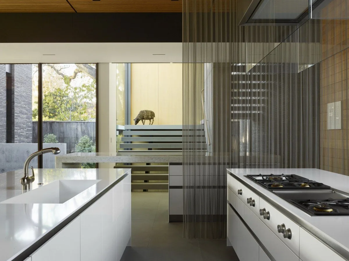 Modern kitchen featuring a white island with integrated sink and gas cooktop, separated by a sheer curtain, with large windows overlooking a stair and outdoor area.