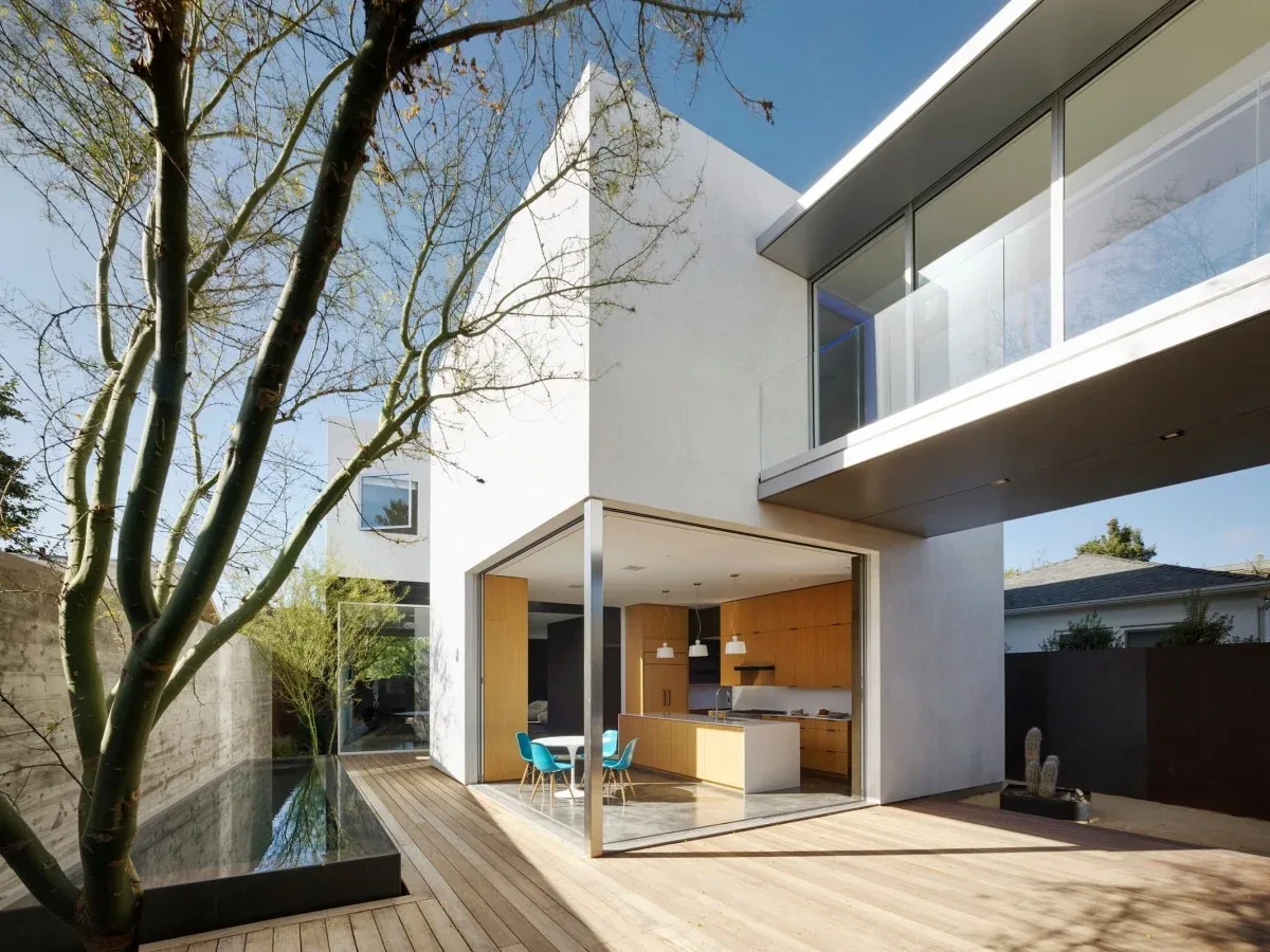 Exterior view of the Spectral Bridge House with an open-plan kitchen and dining area opening to an outdoor wooden deck and reflecting pool.