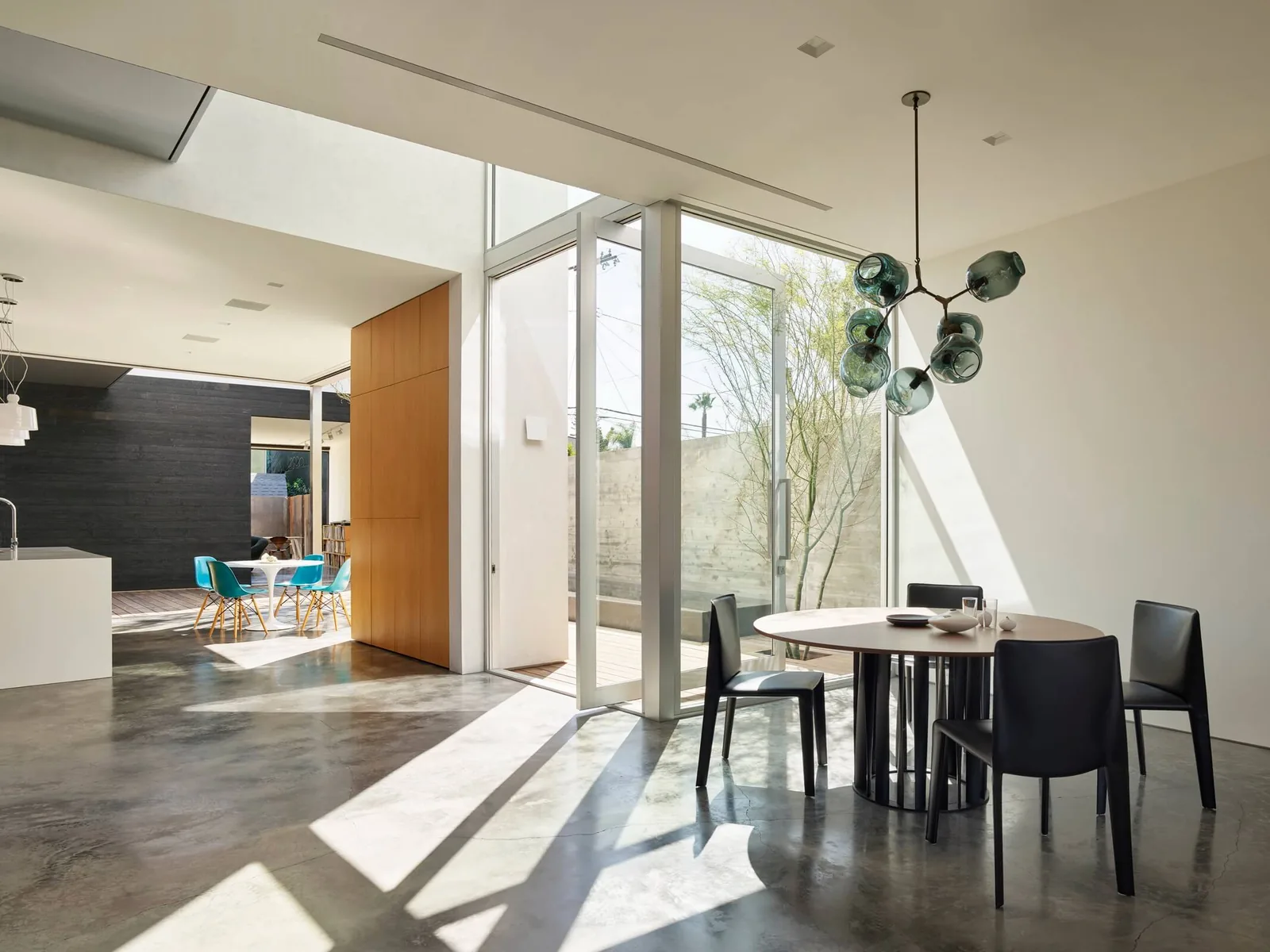 Interior view of a dining room with large glass doors opening to an outdoor space, adjacent to a kitchen and seating area with blue chairs.