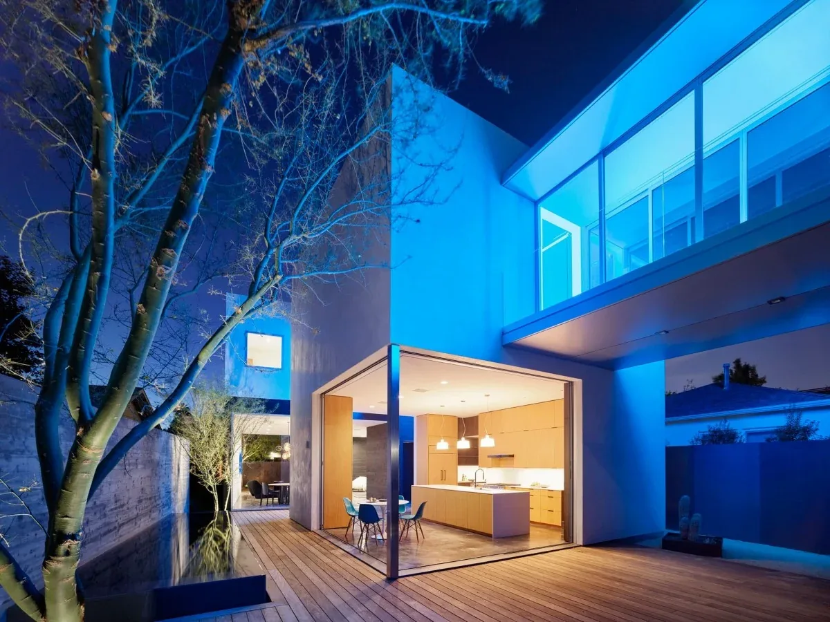 Modern house exterior showing an open kitchen and wooden deck at night with blue lighting.