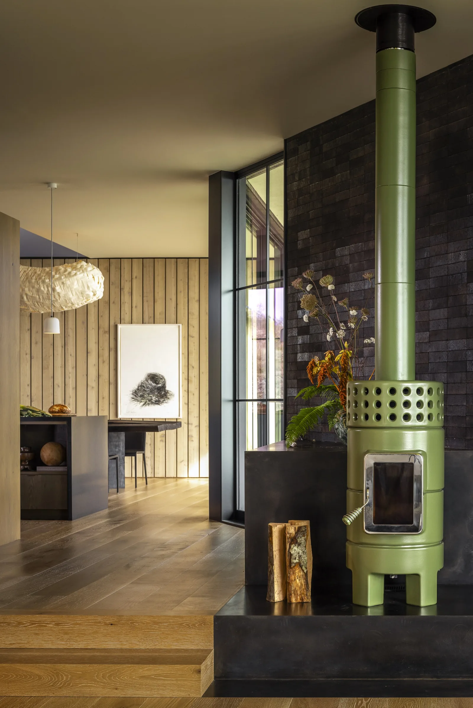Interior view of a wood-paneled room with a green stove and black hearth, featuring a wood floor and a woven pendant light above a dining table.