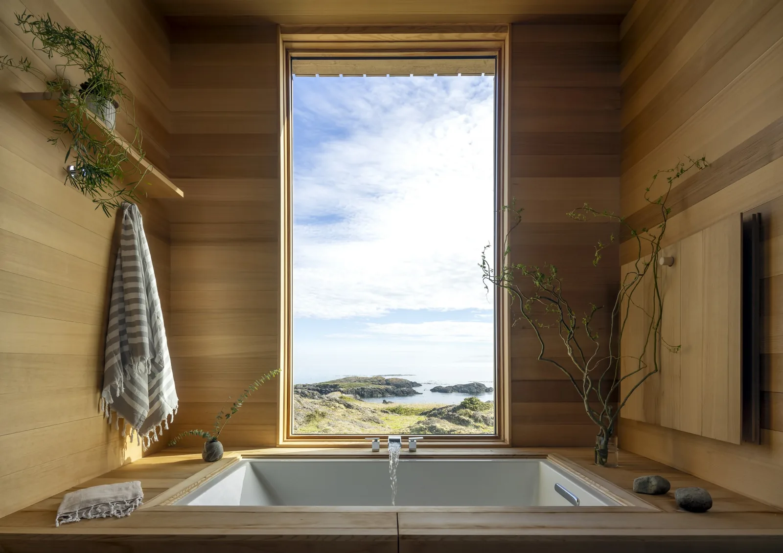 Bathtub with wood paneling walls features a large window framing a rocky coastal landscape in False Bay project.