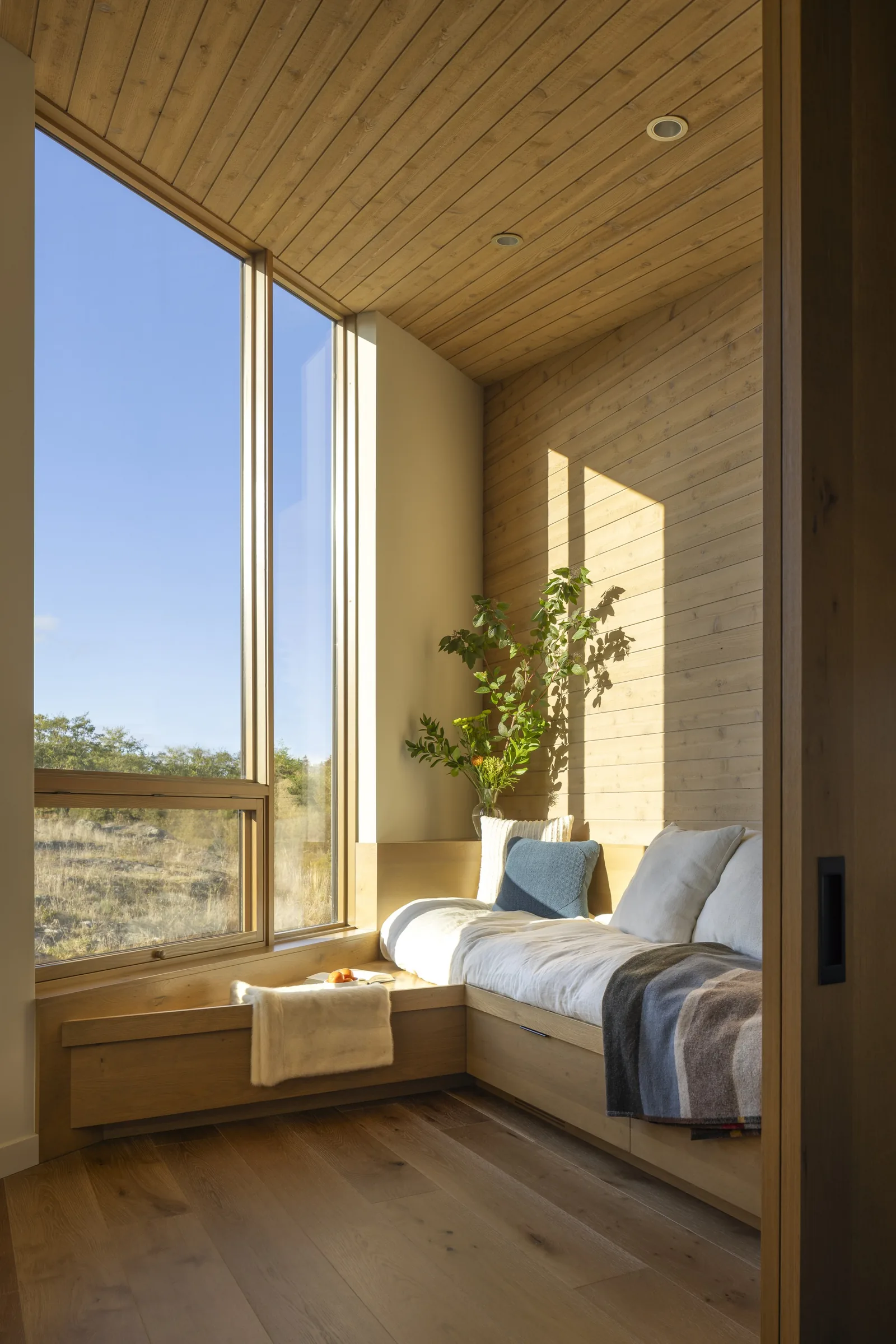 A bright nook with a built-in wooden bench seat is surrounded by floor-to-ceiling windows and wood-paneled walls and ceiling.