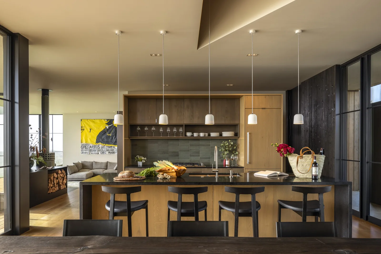 The kitchen features a dark kitchen island with wood cabinetry and pendant lighting, opening to a living space with wood storage and black window frames.
