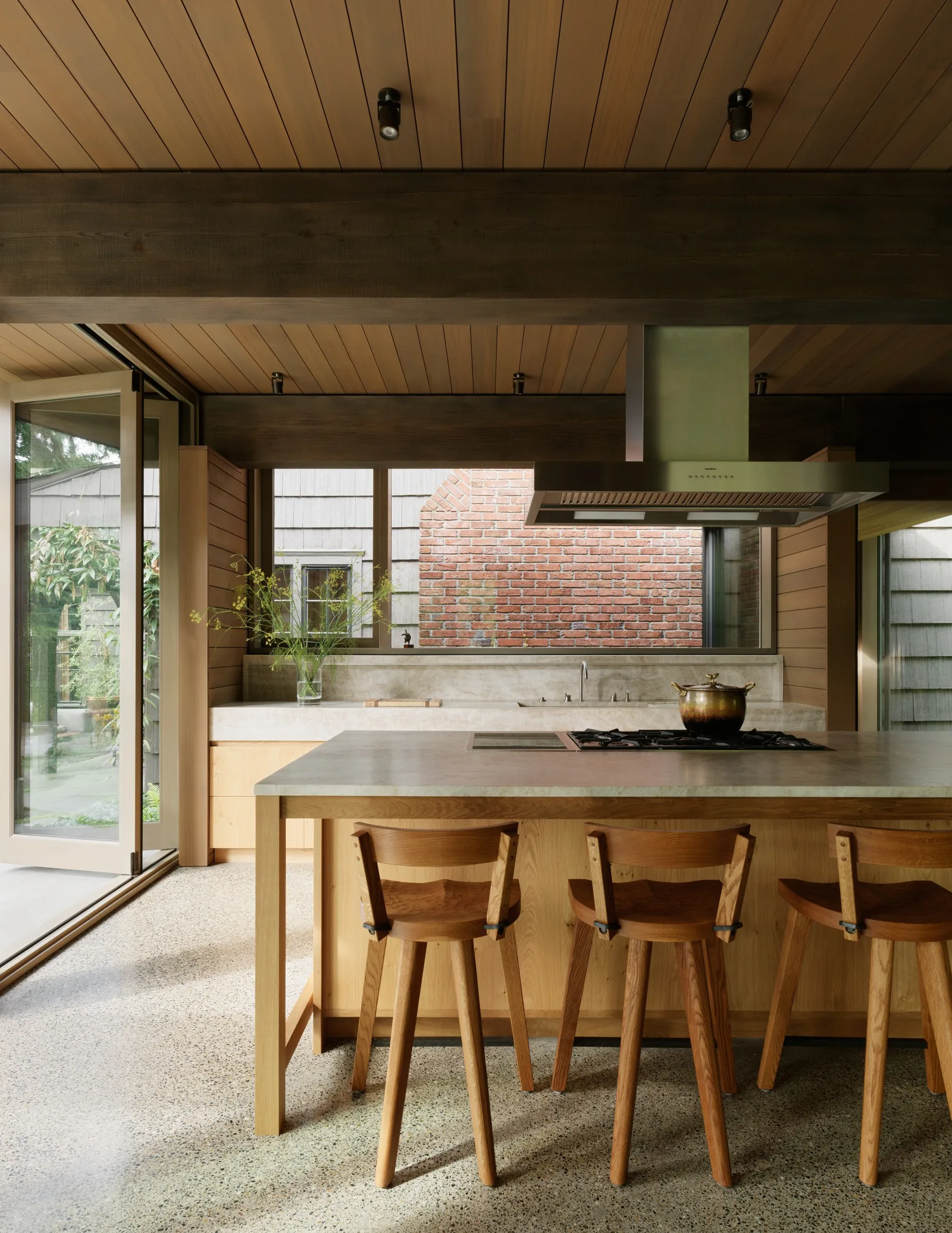 Kitchen interior with wood paneling, concrete countertop island with cooktop, and floor-to-ceiling glass doors opening to an outdoor space.