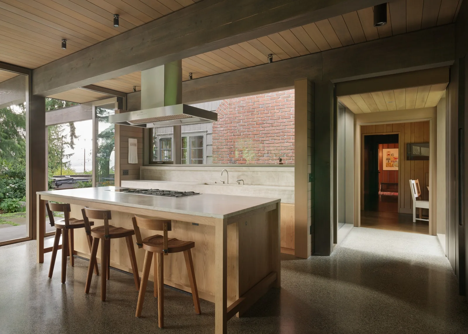Modern kitchen interior with exposed beams, built-in island with cooktop, and large windows looking into a garden.