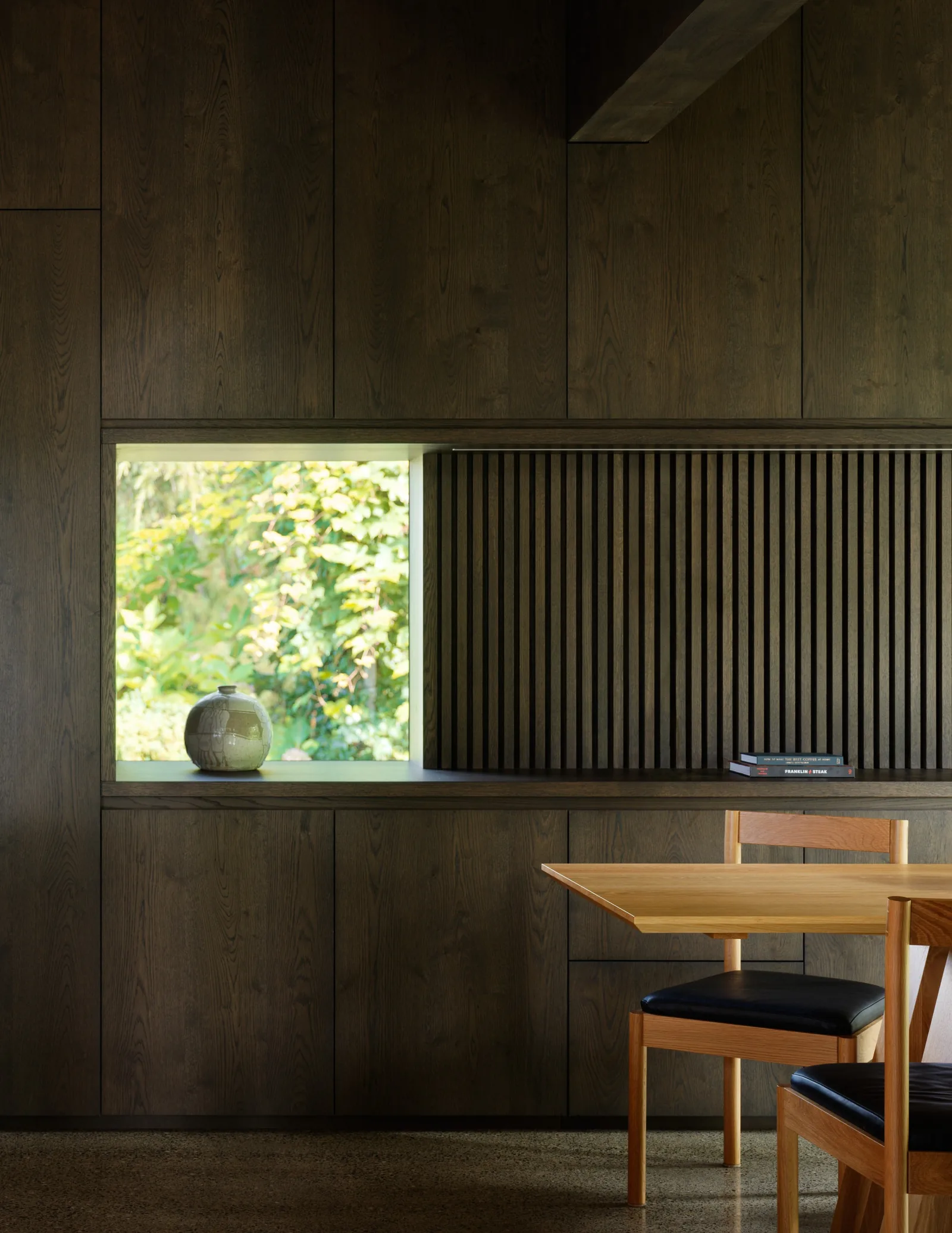 Dark wood-paneled wall features a built-in horizontal window and a vertical slatted wood design above a built-in shelf beside a wood dining table and chairs.