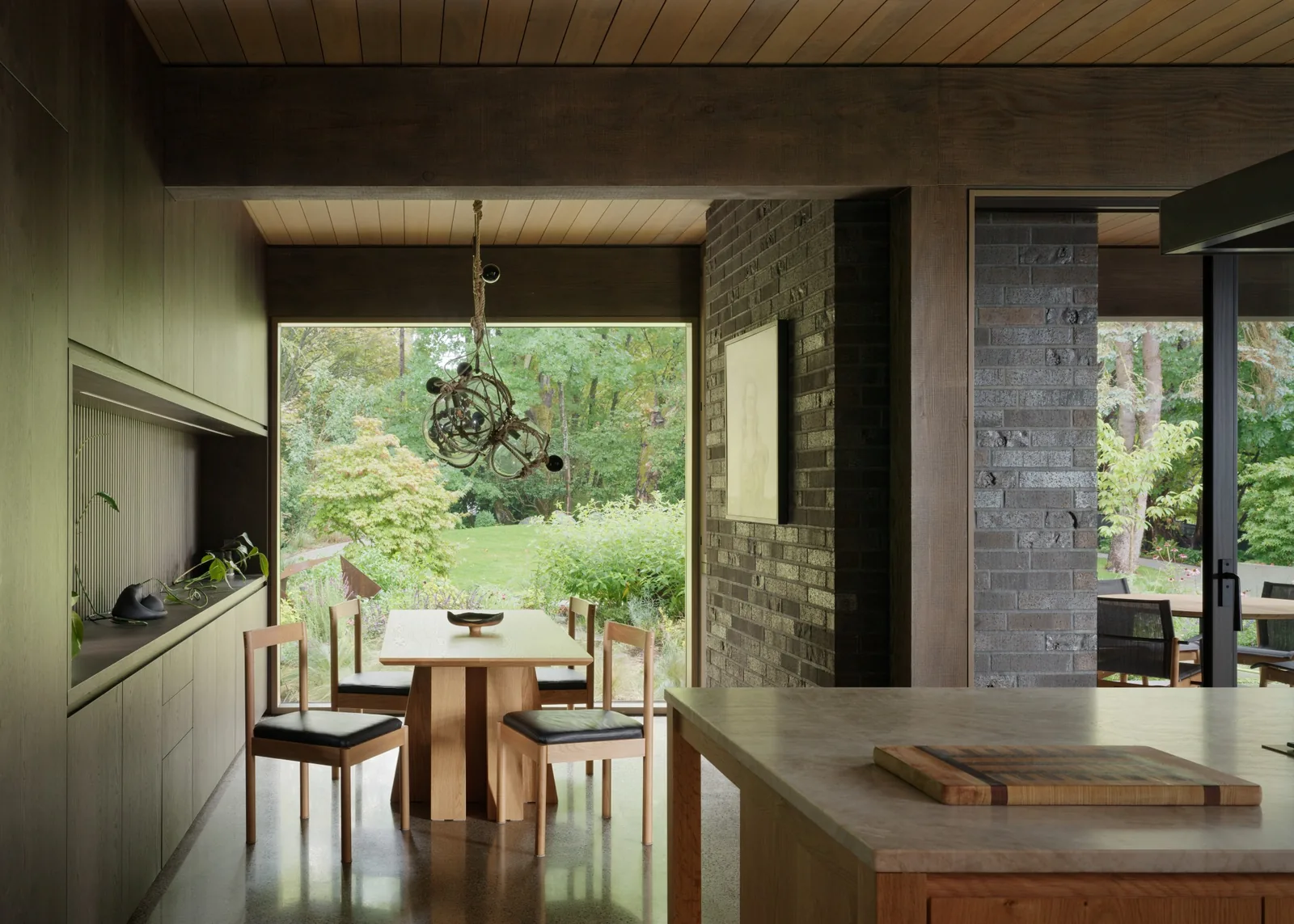 The dining area features a wood table and chairs, a large window overlooking a lush garden, and an adjacent kitchen island.