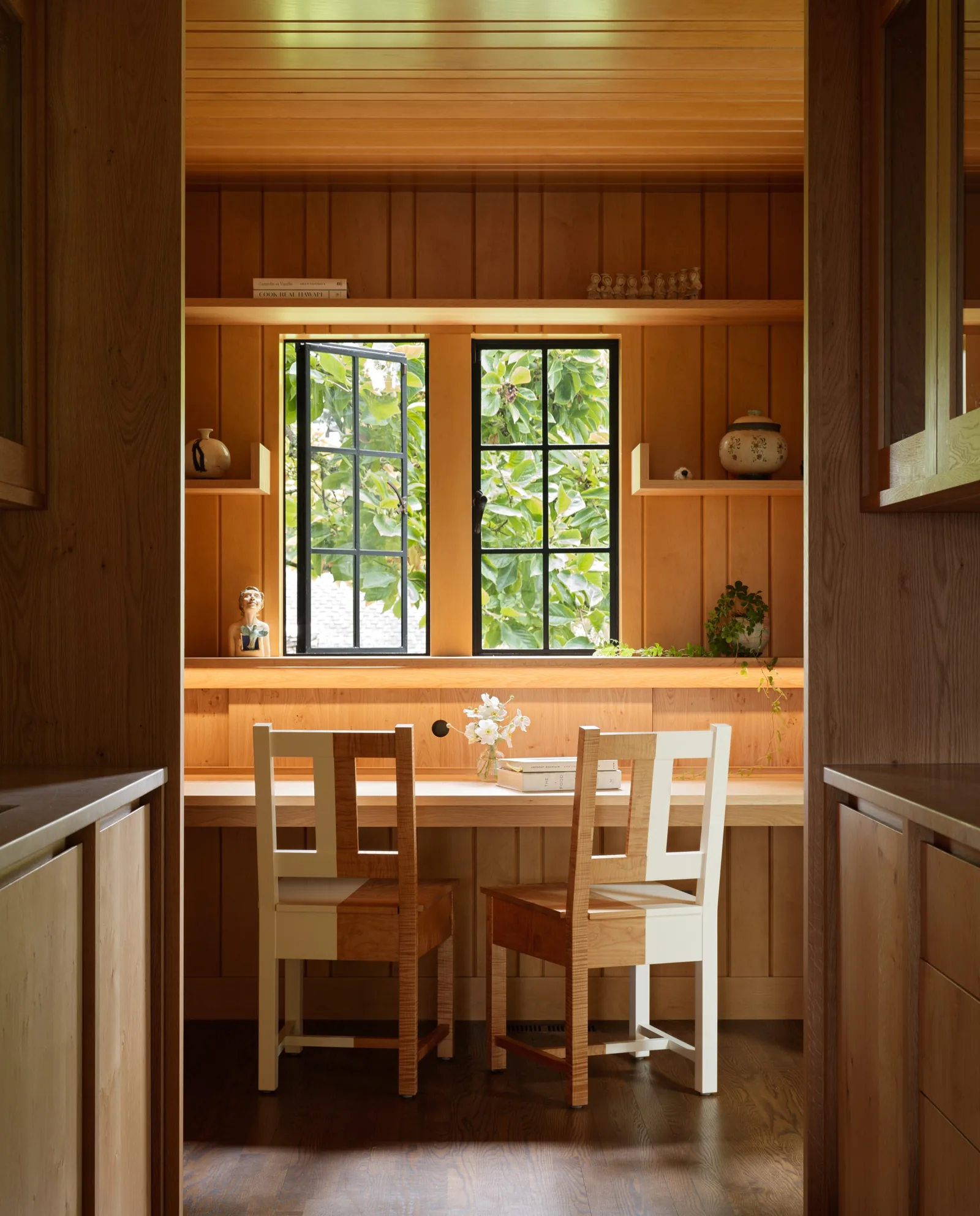 Dining nook with wood paneling, built-in shelves, and black-framed casement windows overlooking greenery.