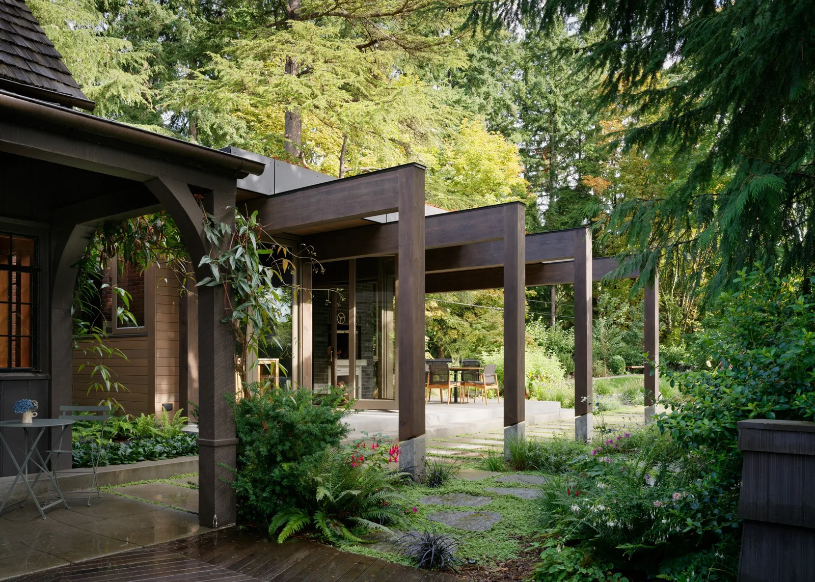 Modern dark wood pergola covering an outdoor patio connected to a house with large glass doors, surrounded by garden vegetation.