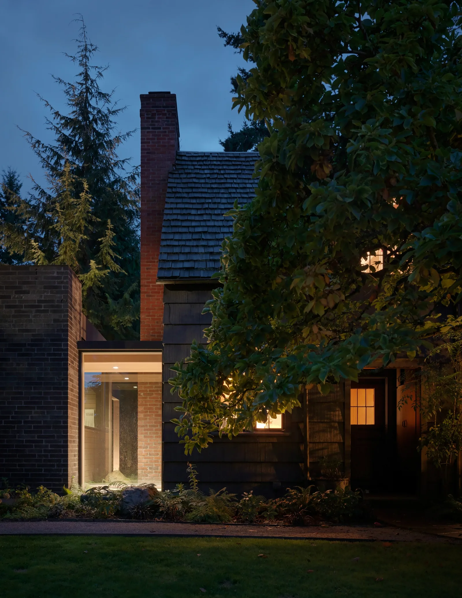 Exterior front elevation of a residential building at dusk with brick chimney and shingle roof.