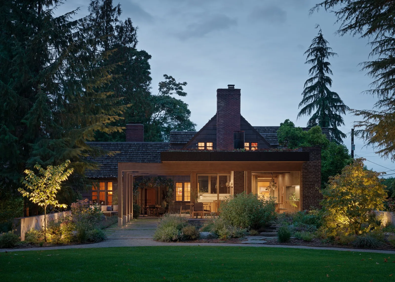 Dusk view of the rear exterior of a brick house featuring a wooden pergola patio and garden landscape.