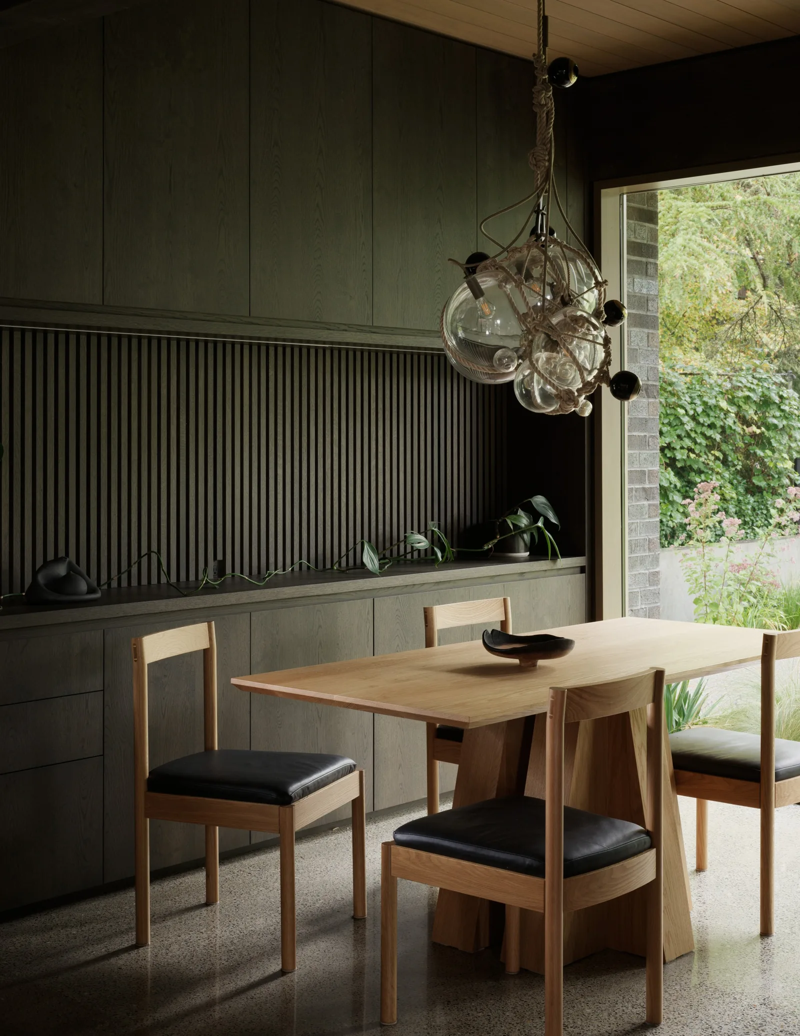 Dining area with a wooden table, black-cushioned chairs, and custom dark wood wall paneling next to a large window overlooking greenery.