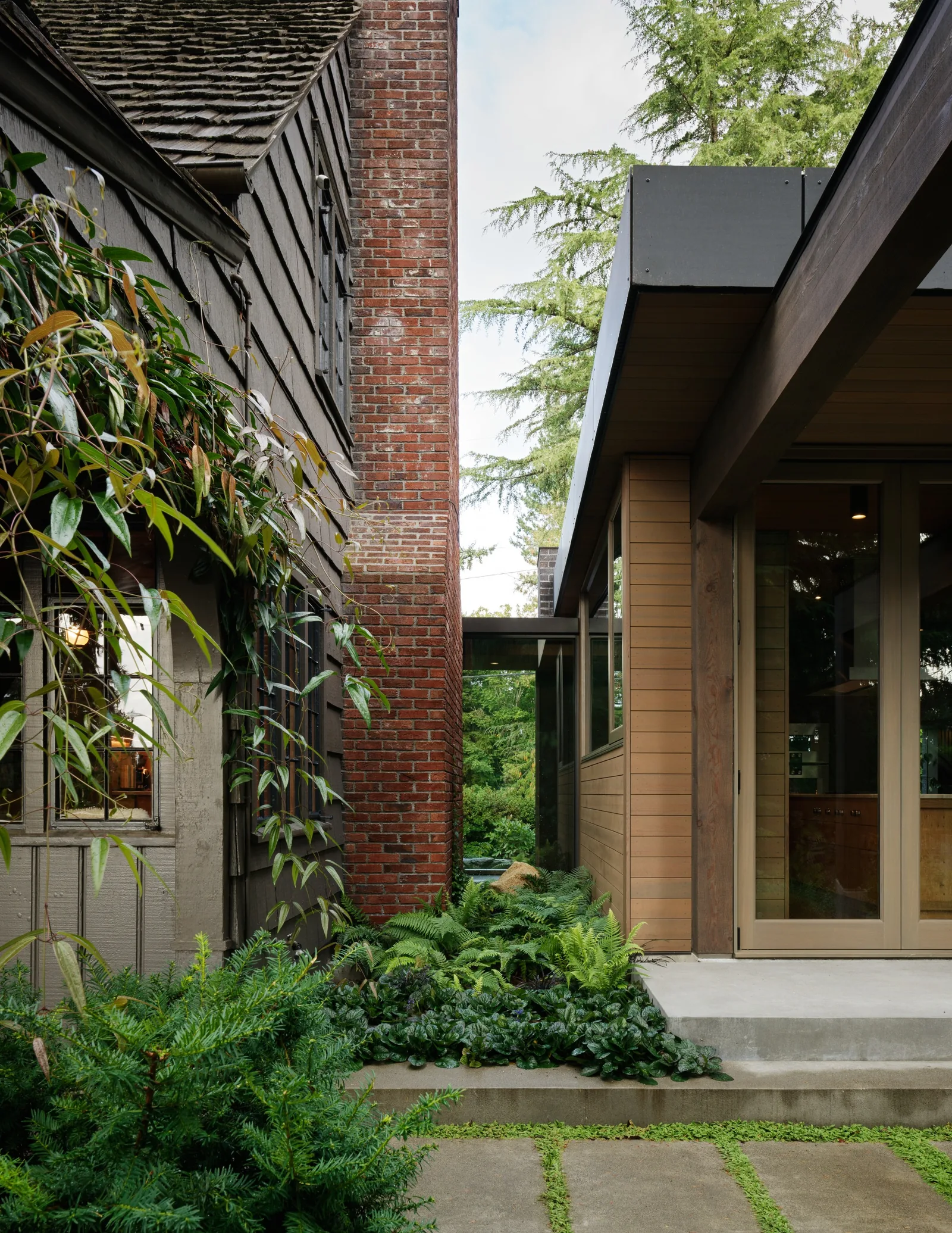 Transition between a traditional house with wood shingle siding and brick chimney and a modern wood-paneled addition with large glass door.
