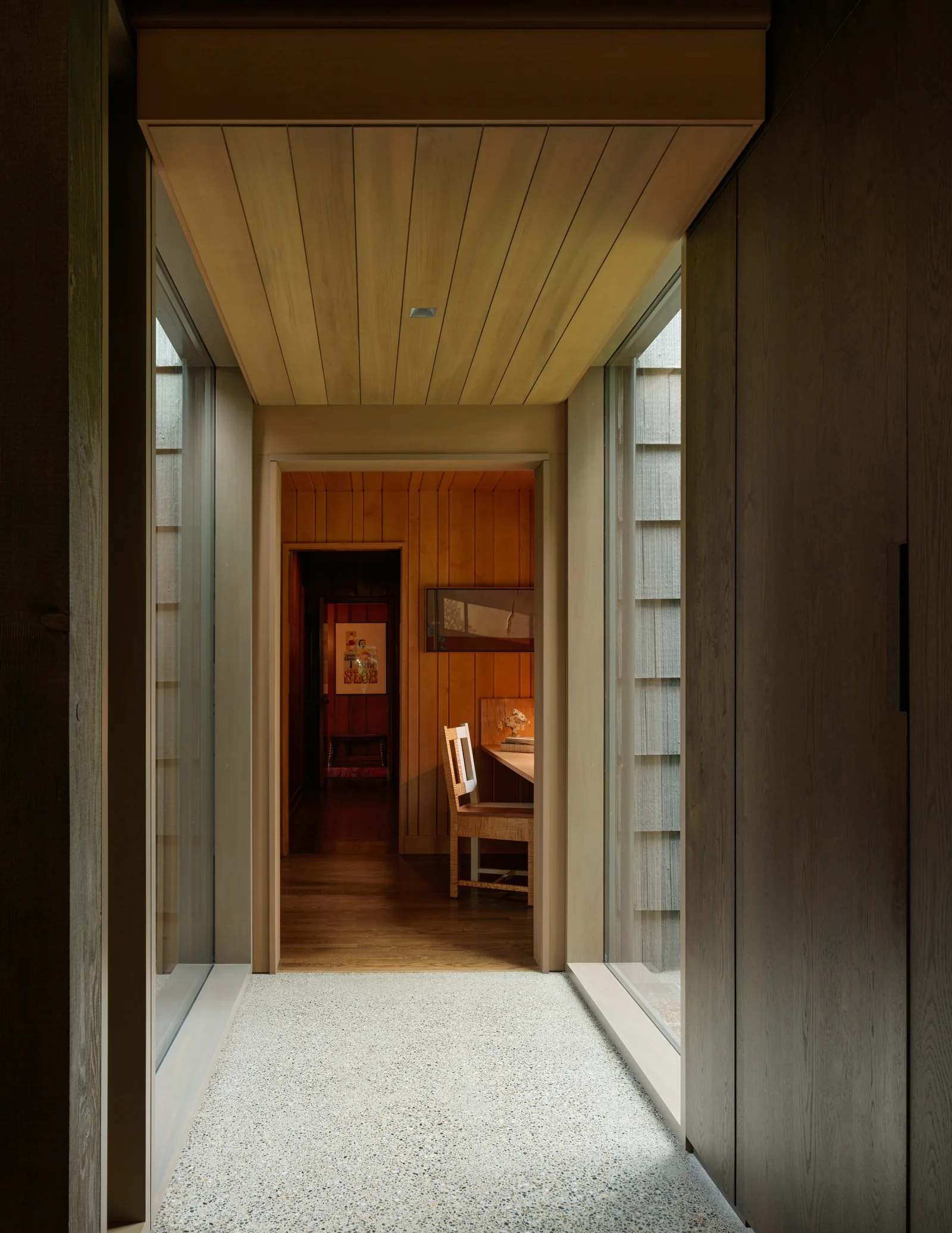 A hallway features floor-to-ceiling glazing and a wooden ceiling leading to a wood-paneled room with a chair and artwork.