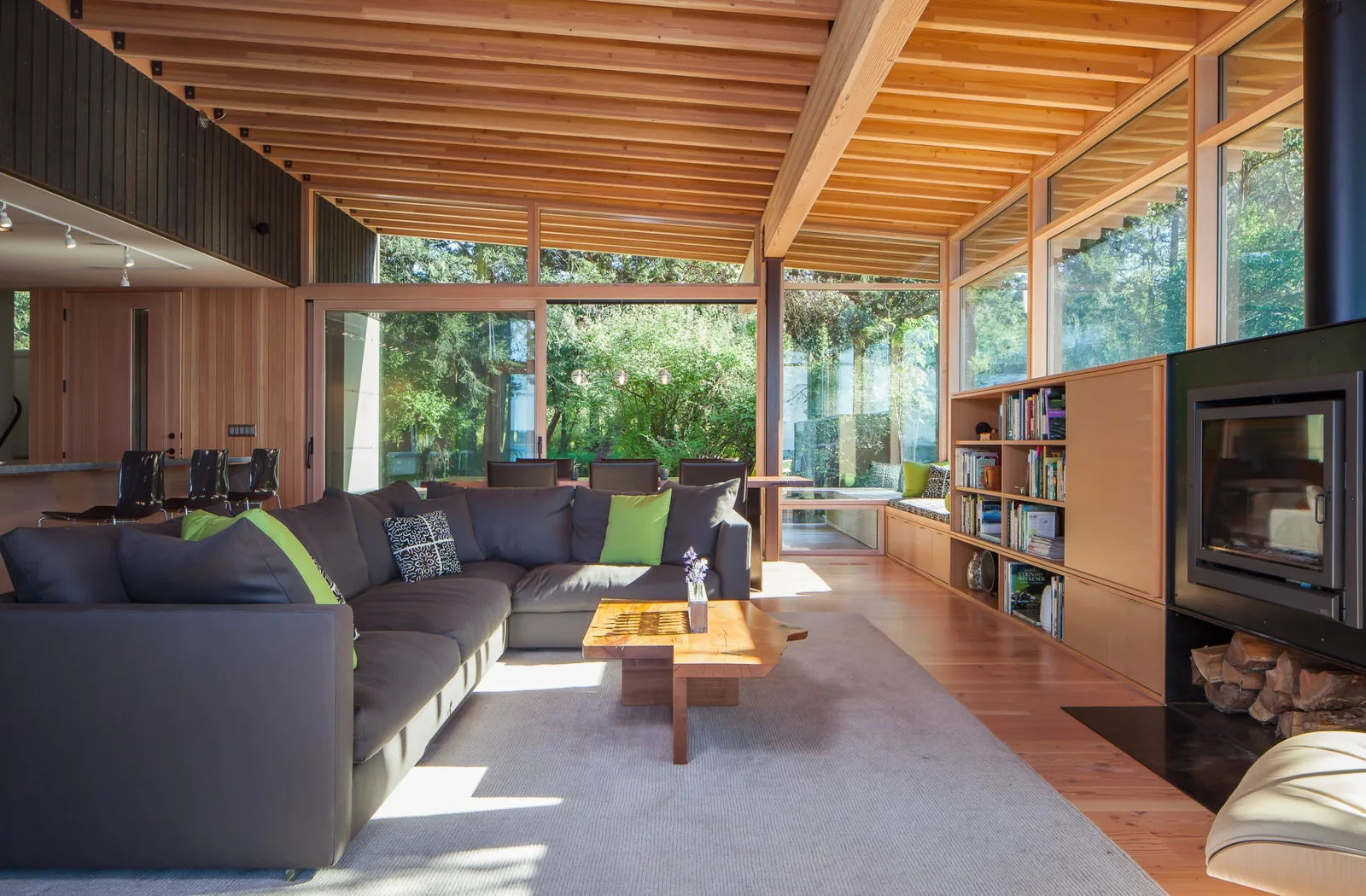 Contemporary living room with exposed wood ceiling beams, built-in shelving, and large windows overlooking greenery.