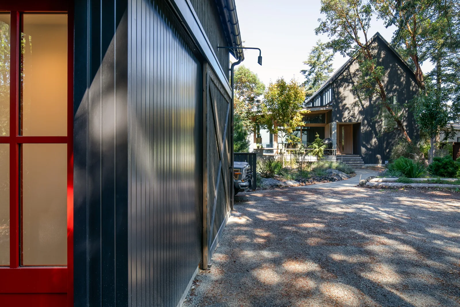 Side exterior view showing dark vertical siding, red-framed frosted glass door, gravel driveway, landscaping, and a gable roof house in background.