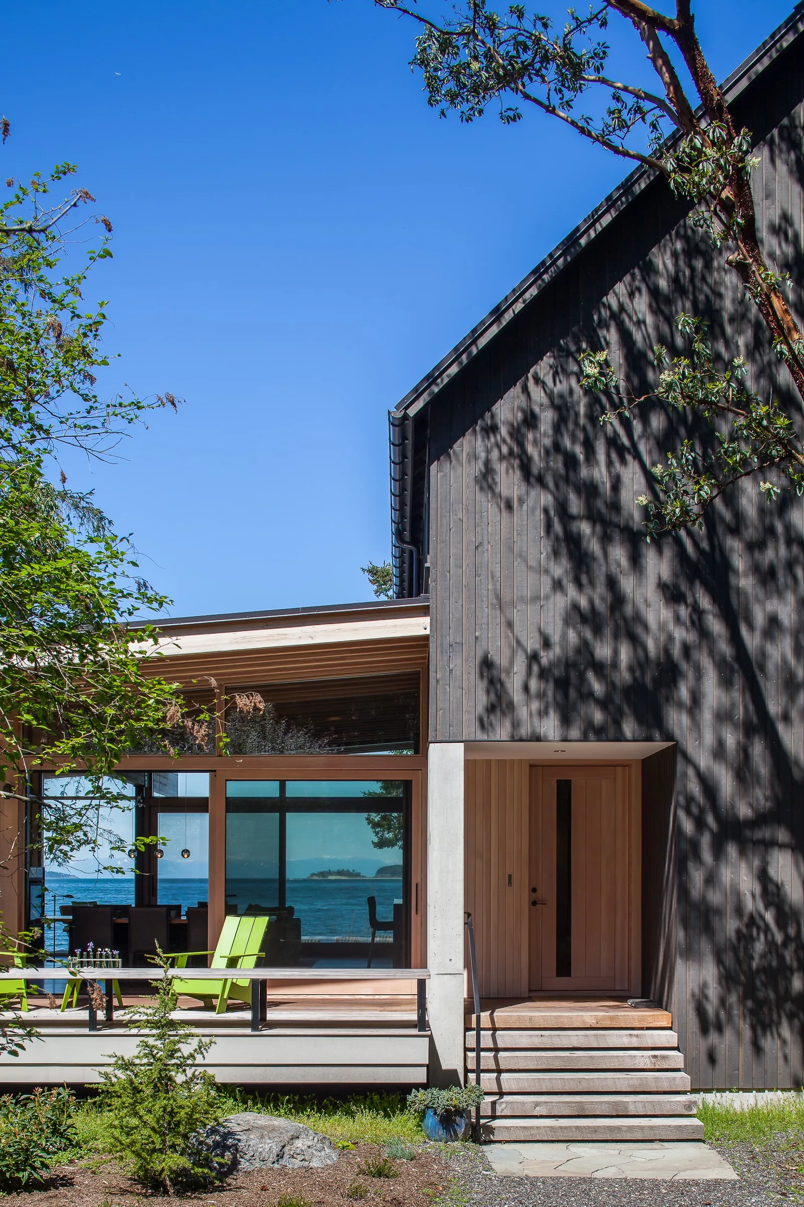 Modern house exterior with dark vertical wood siding and light wood entry door under a clear blue sky.