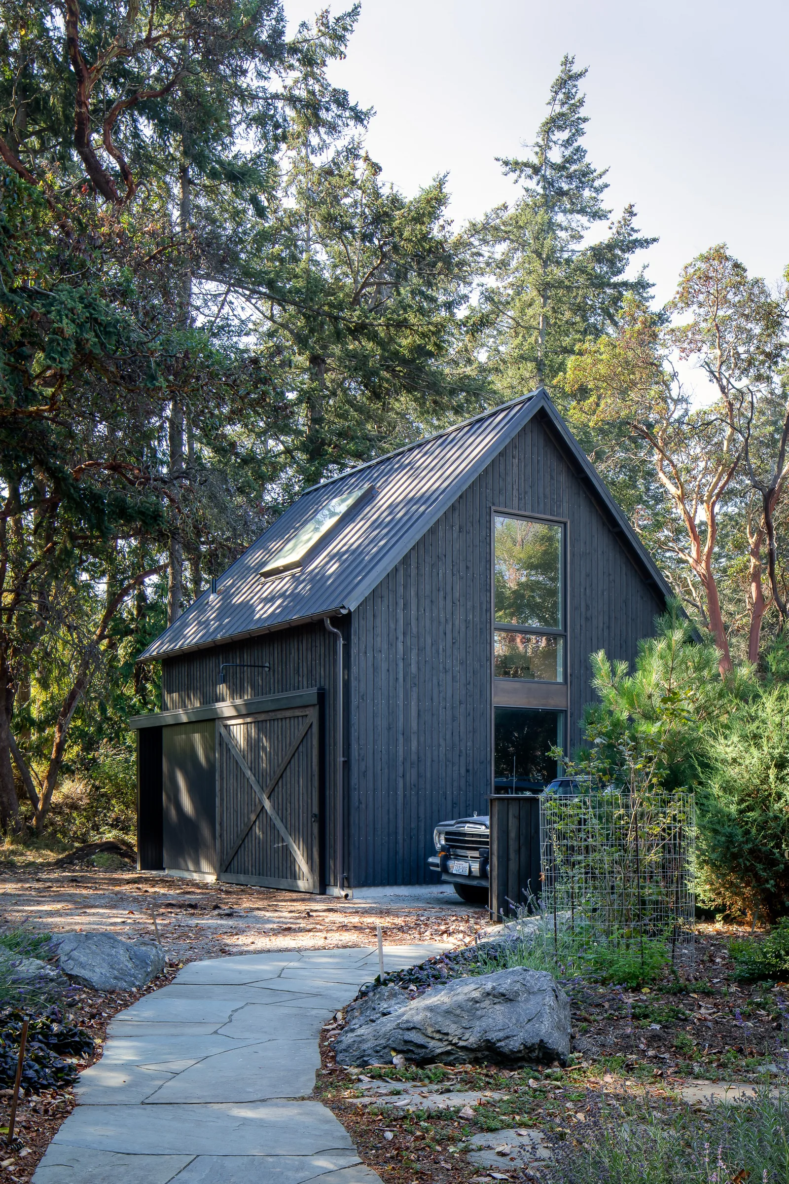 A detached dark gray garage with a gable roof and a large vertical window nestled within a wooded landscape.