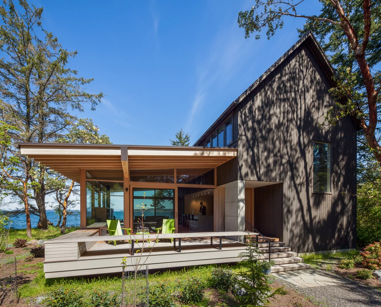 Modern house exterior showing dark vertical wood siding, gable roof, and covered patio with floor-to-ceiling windows.
