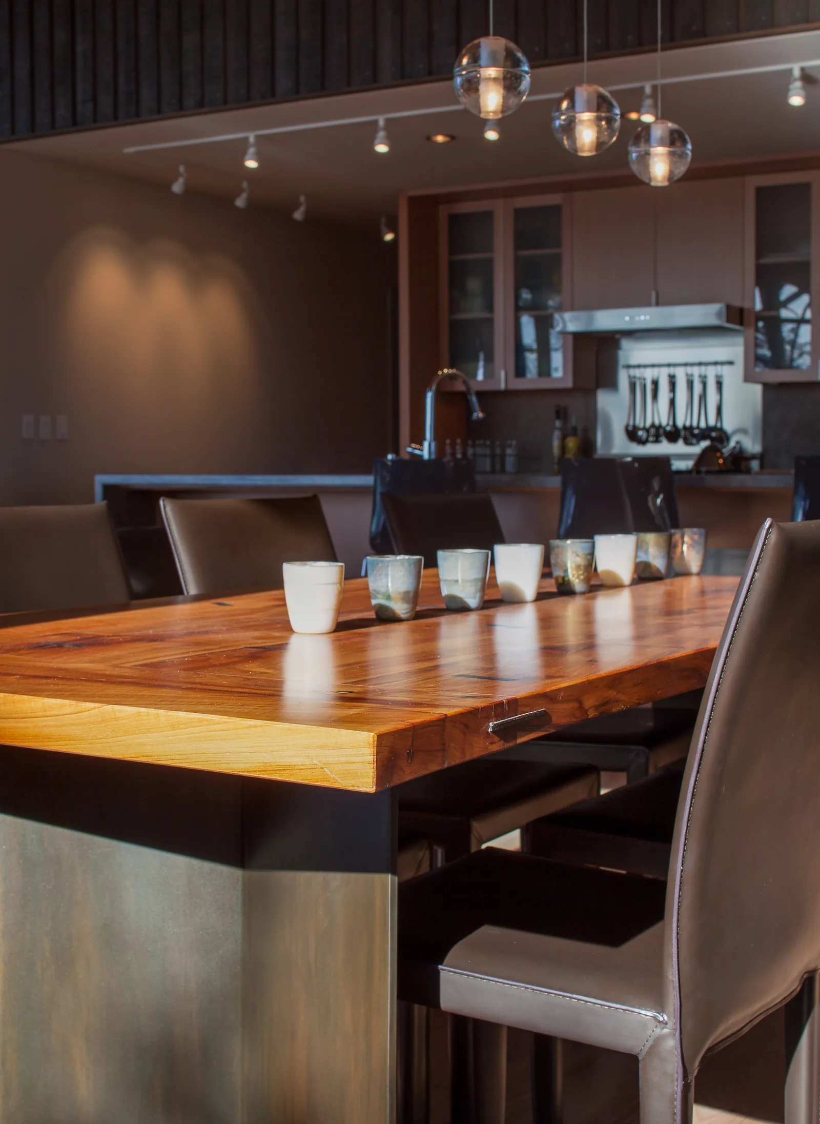 Kitchen with a wood dining table, leather chairs, pendant lighting, and a cooking area with cabinets and stove hood in the background.